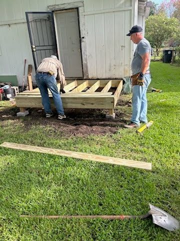 Two volunteers framing the landing of a new ramp at the back door of a white house, floor joists in place and lumber lying in the grass.